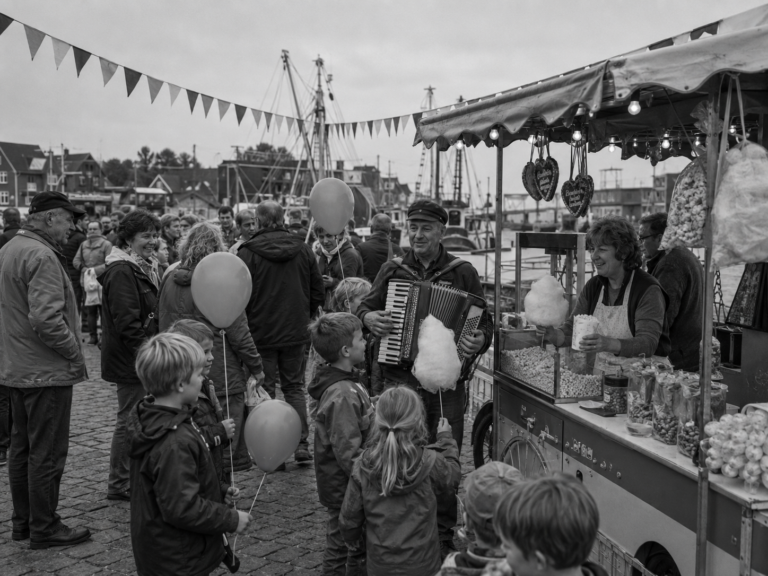 Schwarzweißaufnahme eines Hafenfestes mit Besuchern, Kindern, Luftballons, Fischstand und Akkordeonspieler.