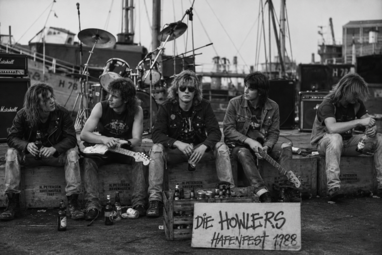 Schwarzweißaufnahme von fünf Jugendlichen der Band „Die Howlers“, die beim Hafenfest 1988 auf Fischkisten hinter einer improvisierten Bühne am Hafen sitzen; mit Gitarren, Verstärkern und Getränkeflaschen.